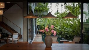 Uma área de refeições com mesa e cadeiras de madeira, um vaso com flores cor-de-rosa e plantas pendentes.