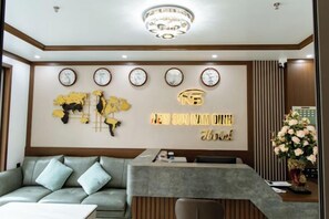 Een receptiegedeelte met een bank, een receptiebalie, een wandklok en een decoratief wandobject.