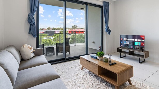 A modern living room with a grey sofa, a wooden coffee table, a flat-screen TV, and a sliding glass door leading to an outdoor area with a view of a red building and greenery. - 2-Bedroom Apartment Balcony Close to All Hotspots (MacGregor)