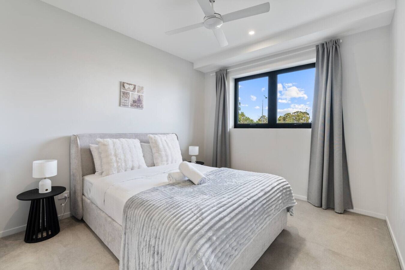 A bedroom with a bed, a ceiling fan, a window with curtains, and a small table with a lamp.