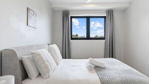 A modern hotel room with a large bed, grey curtains, a ceiling fan, and a window with a view of the outdoors. - 2-Bedroom Apartment Balcony Close to All Hotspots (MacGregor)
