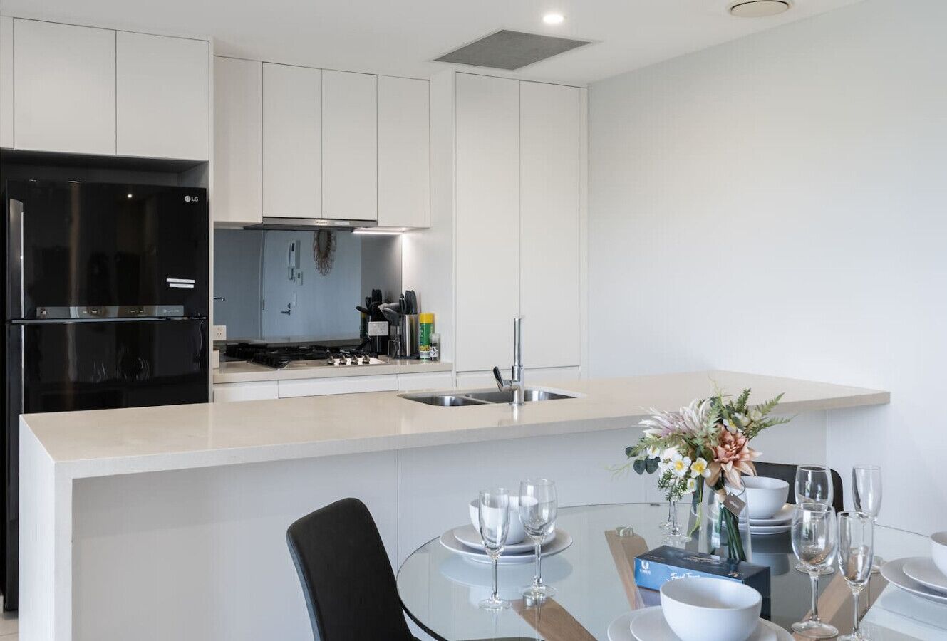 A modern kitchen with white cabinetry, a black refrigerator, and a dining area with a glass table set for two.