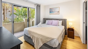A bedroom with a bed, a nightstand, a window with a view of greenery, and a painting on the wall. - Spacious 2BR Getaway | Great Location & Comfort (Indooroopilly)