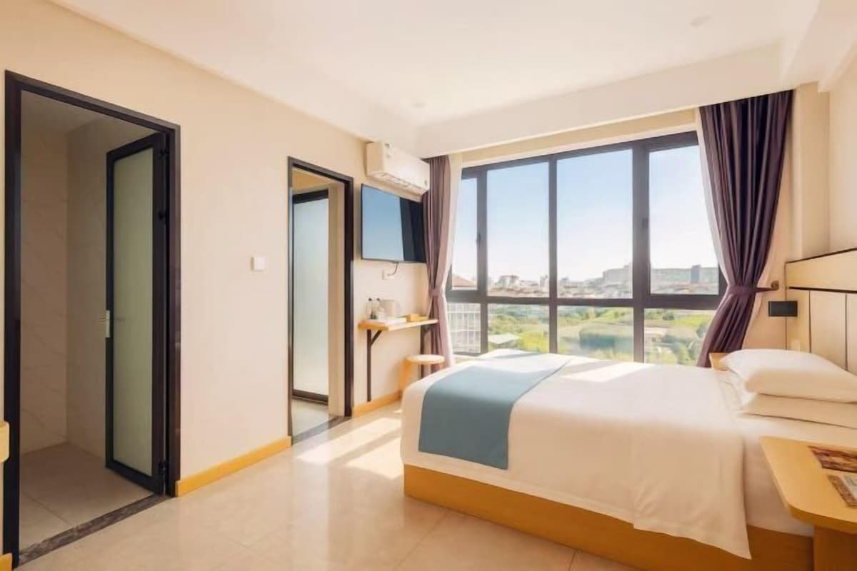 A hotel room with a bed, a desk, a chair, a window with a city view, and a door leading to another room.