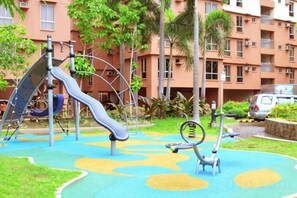 A playground with slides and swings, surrounded by a green lawn and a building with balconies. - Near Manila Airport. Lovely "HOTEL STYLE" Studio Condo, With Kitchenette + Swimming Pool, 7A (Paranaque)