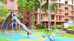 Un parque infantil con toboganes y columpios, rodeado de césped y un edificio con balcones.