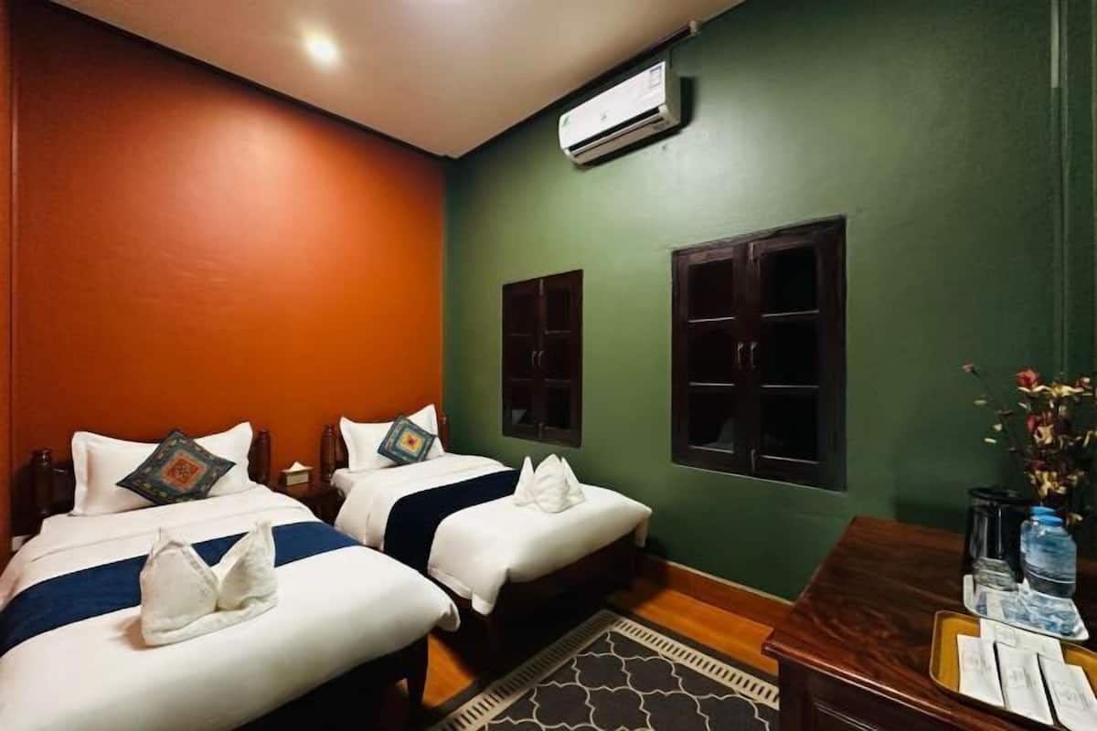 A hotel room with two beds, a wooden headboard, an air conditioner, a desk with a water bottle, and a vase with flowers.