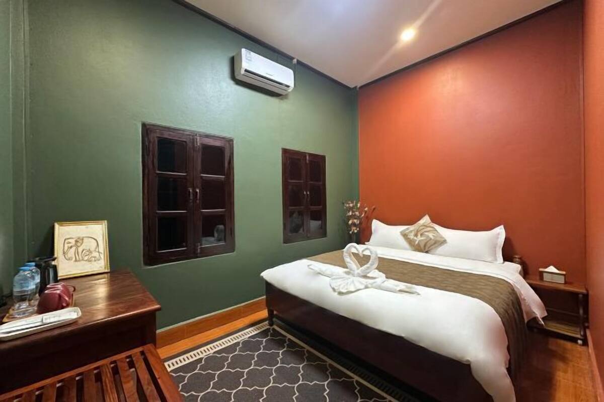 A hotel room with a bed, a desk with a chair, an air conditioner, and two windows with wooden frames.