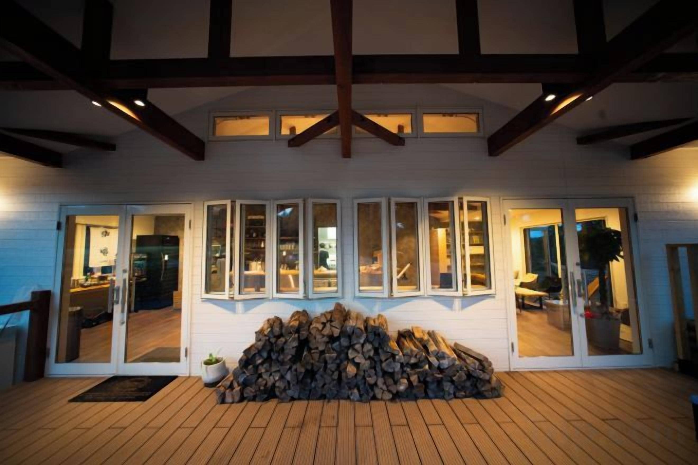 A covered outdoor area with a stack of firewood, a wooden deck, and a building with glass doors and windows.