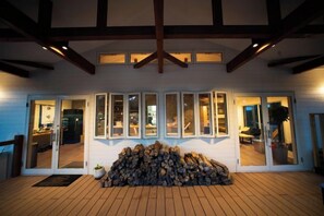 A covered outdoor area with a stack of firewood, a wooden deck, and a building with glass doors and windows.