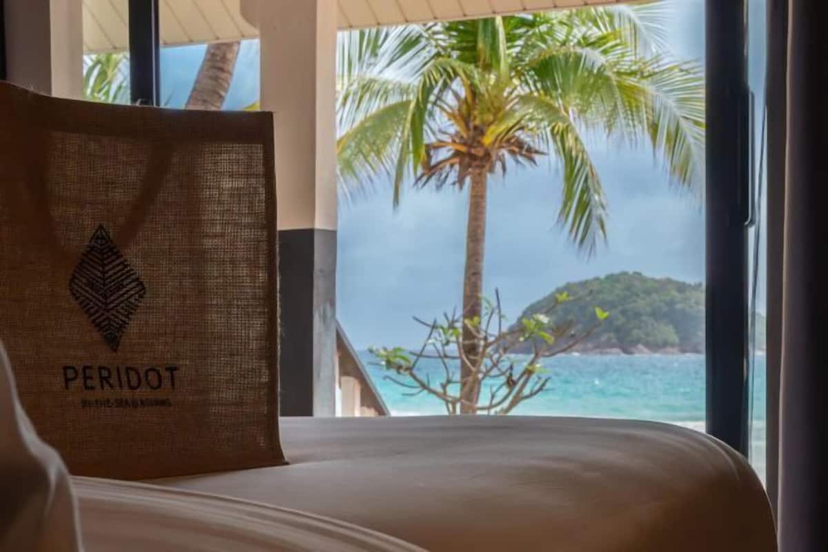A bed with a pillow labeled 'PERIDOT' and a view of the ocean and palm trees.
