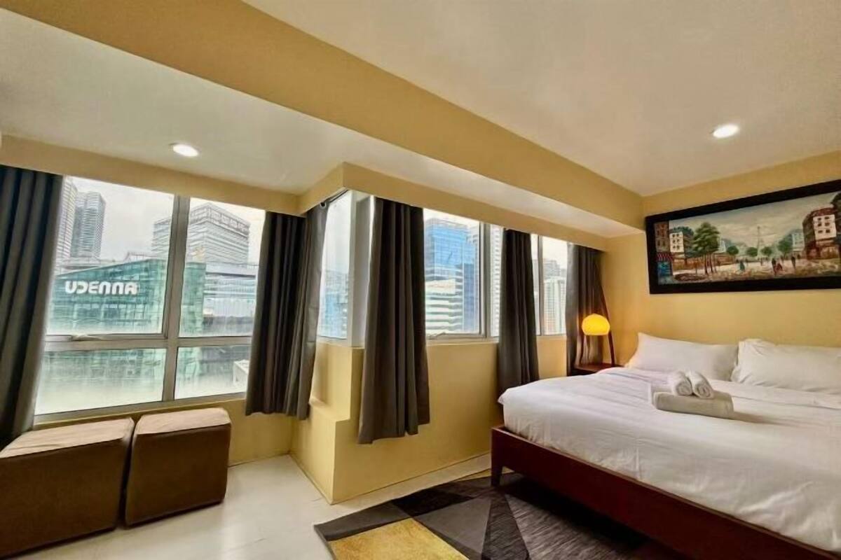 A hotel room with a large window, a bed, a bedside table with a lamp, and a painting on the wall.