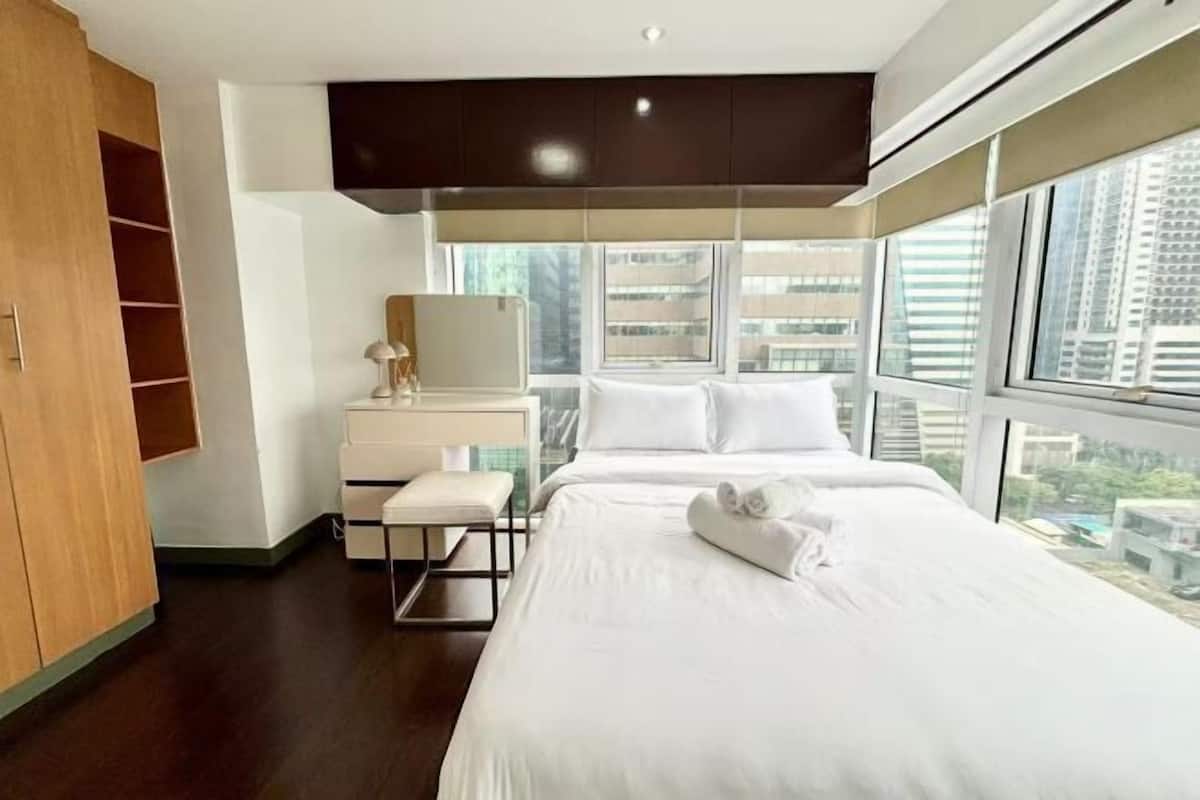 A modern hotel room with a large bed, a desk with a lamp, and a view of the city through large windows.