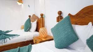 Camera d'albergo con due letti, entrambi con lenzuola bianche e cuscini color acquamarina. Un letto ha una testata in legno e l'altro un comodino di legno con un cesto di vimini.