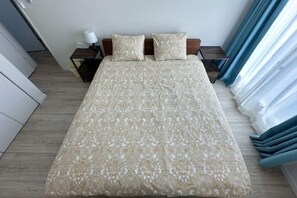 Uma cama bem arrumada com colcha estampada, uma mesinha de cabeceira com abajur e vista para o exterior através das cortinas.