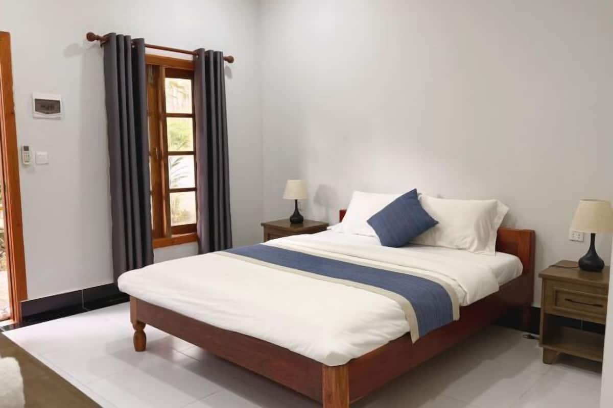 A neatly made bed with a wooden frame, two bedside tables with lamps, a window with curtains, and a thermostat on the wall.
