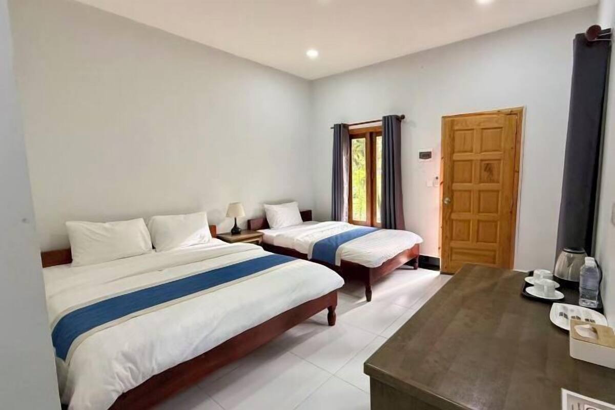 A hotel room with two beds, a wooden door, a small table with a kettle, and a window with curtains.