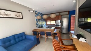 Ein moderner Wohnbereich mit einem blauen Sofa, einem Holzesstisch und Stühlen. Eine Küchenzeile mit Kühlschrank und einem geometrischen Wanddesign ist sichtbar.