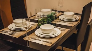 Mesa de comedor dispuesta con platos blancos, tazones, cristalería verde, cubiertos y copas de vino.