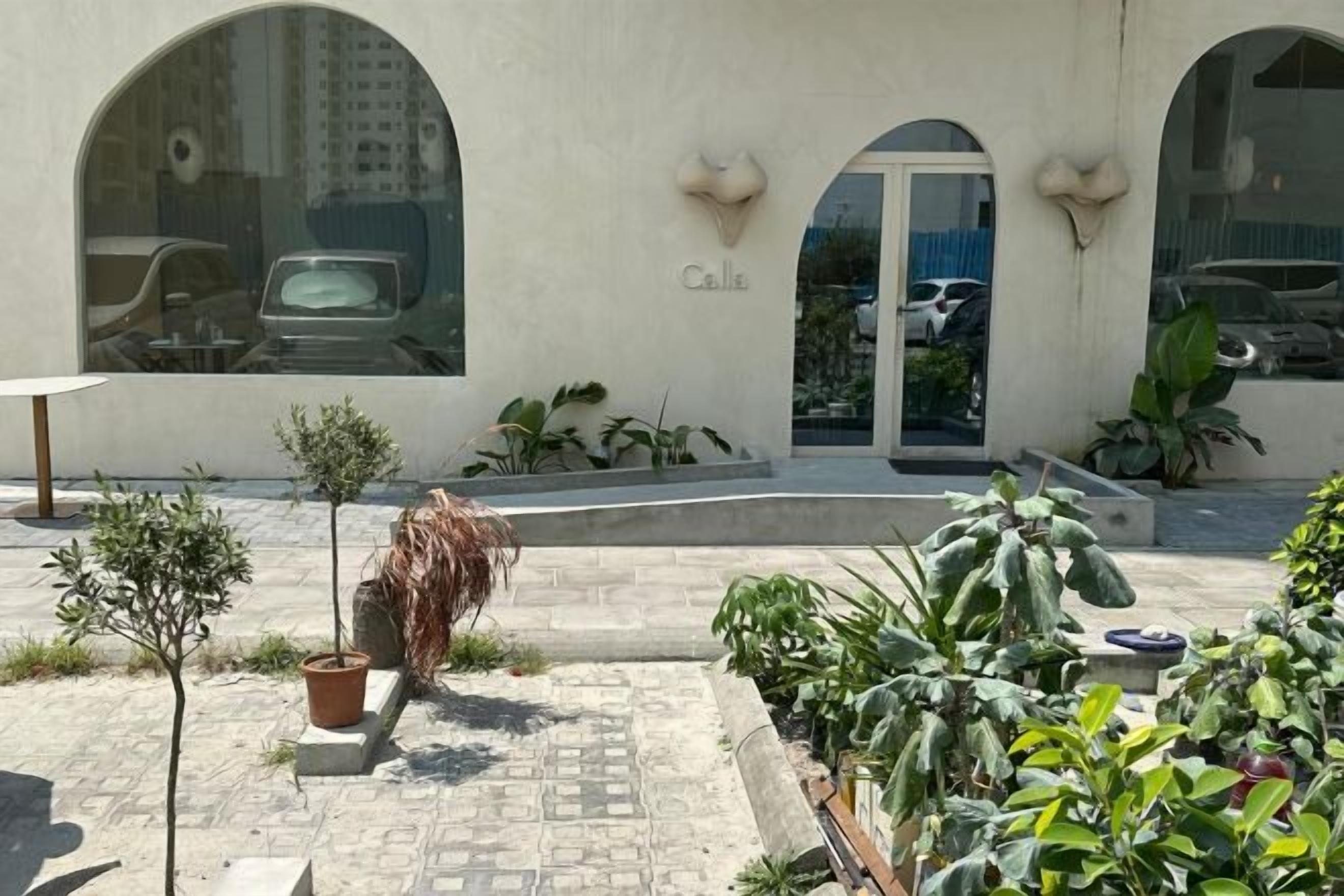 Un edificio con ventanas arqueadas y un letrero que dice 'Calle'. Hay una zona pavimentada con plantas en macetas y una pequeña mesa.