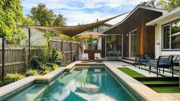 A rectangular swimming pool with a wooden deck and a covered seating area with chairs and an umbrella. - Modern Austin Home w/ Pool, Putting Green & Coffee Nook Townes Retreat by Avantstay (Austin)