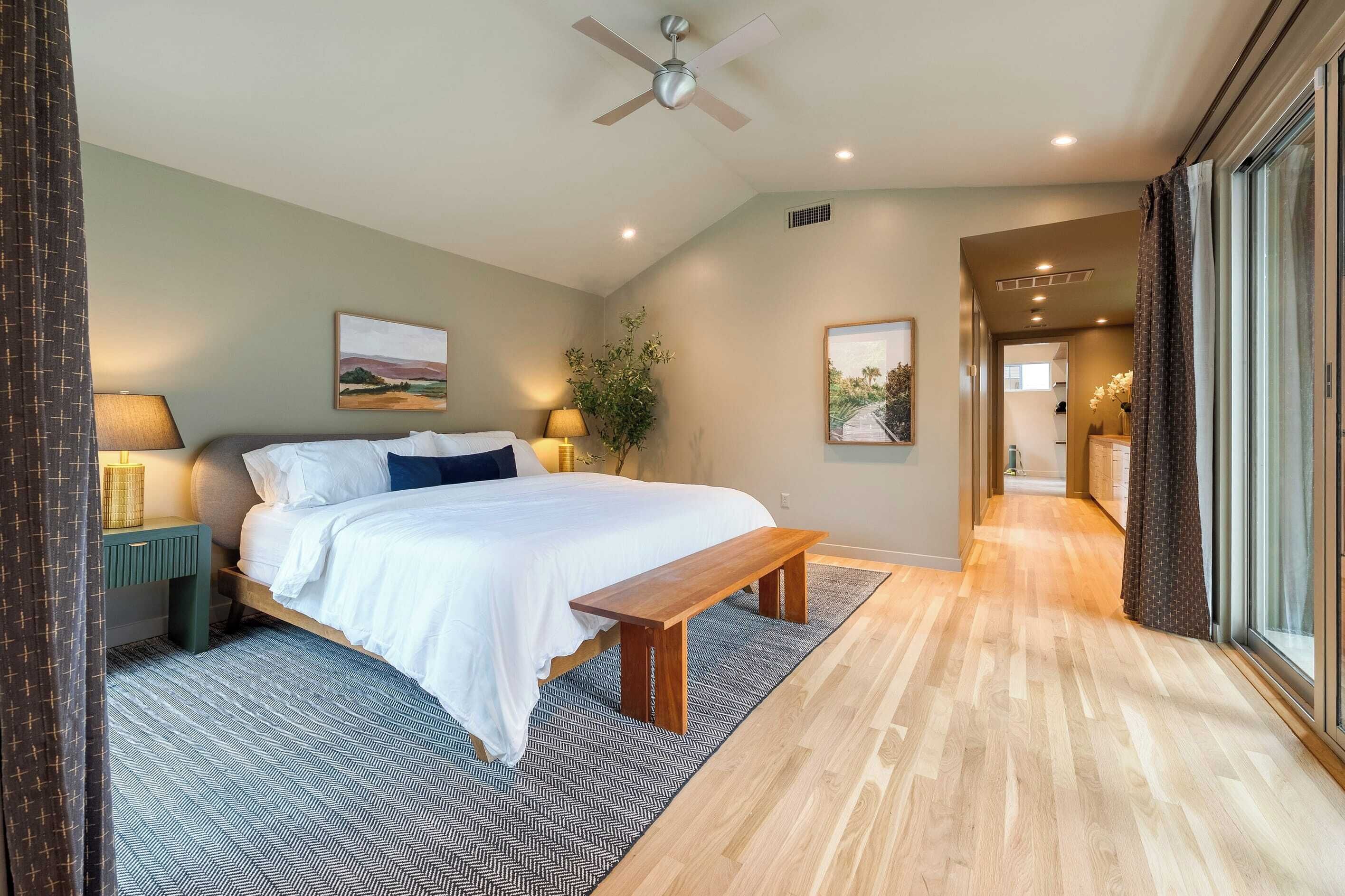 A modern hotel room with a large bed, a wooden bench, a ceiling fan, and a view of the hallway.