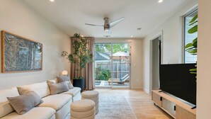 A modern living room with a sofa, ottomans, a TV, and a ceiling fan. - Modern Austin Home w/ Pool, Putting Green & Coffee Nook Townes Retreat by Avantstay (Austin)
