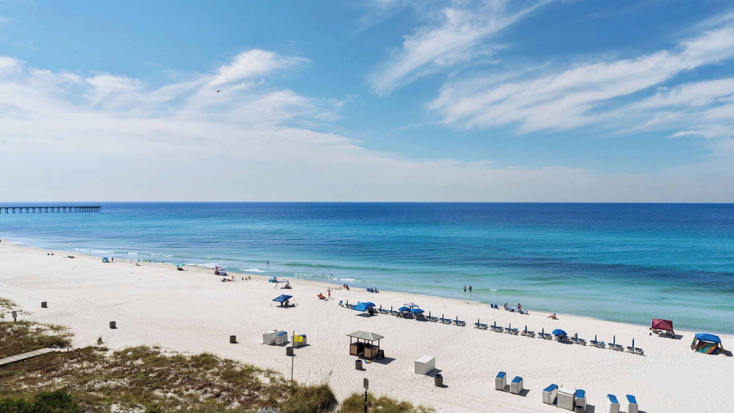 A beach with a pier extending into the water, clear skies, and a sandy shore with beach chairs and umbrellas.