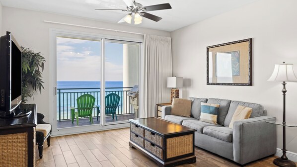 A living room with a grey sofa, a coffee table, a TV, a ceiling fan, a balcony with chairs, and a view of the ocean. - Beachfront Stay w/ Resort Amenities & Gulf Views Sterling Breezing #506 (Panama City Beach)