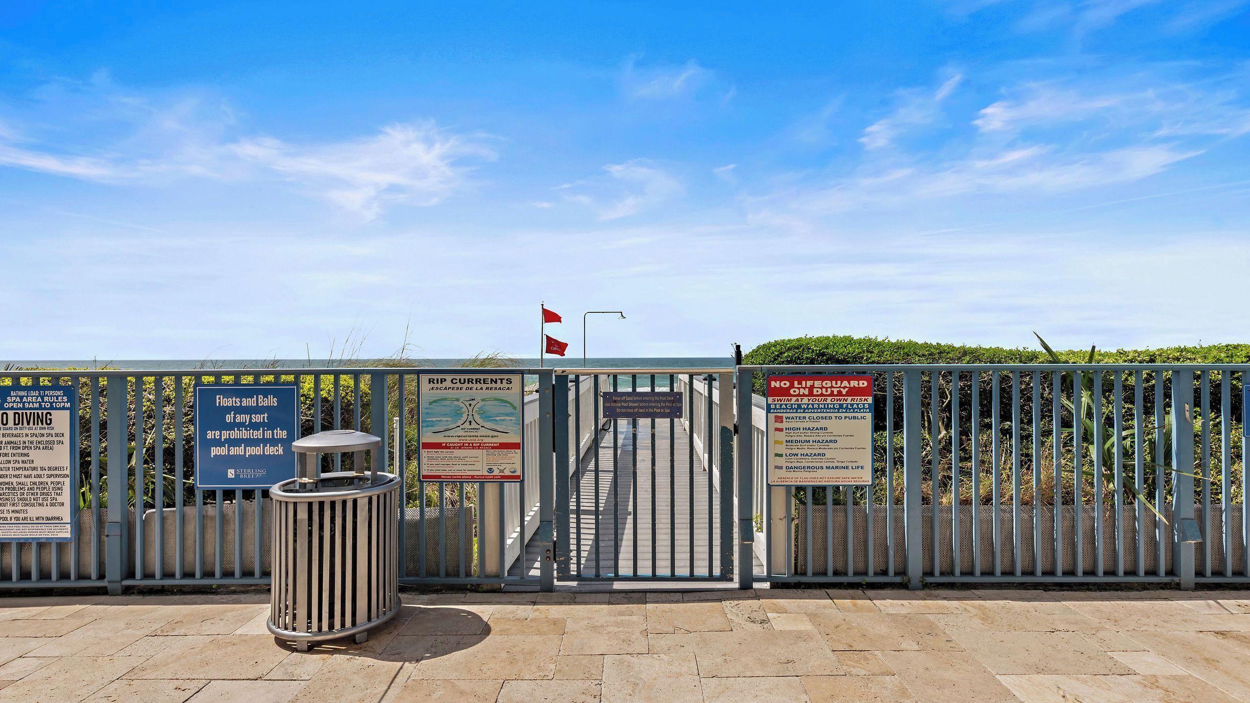 A gated entrance to a pool area with multiple warning signs, a trash can, and a flag.