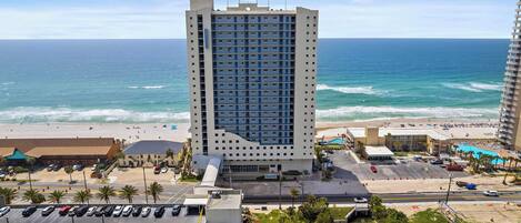 A high-rise hotel with multiple floors, a parking lot, and a beach in the background.