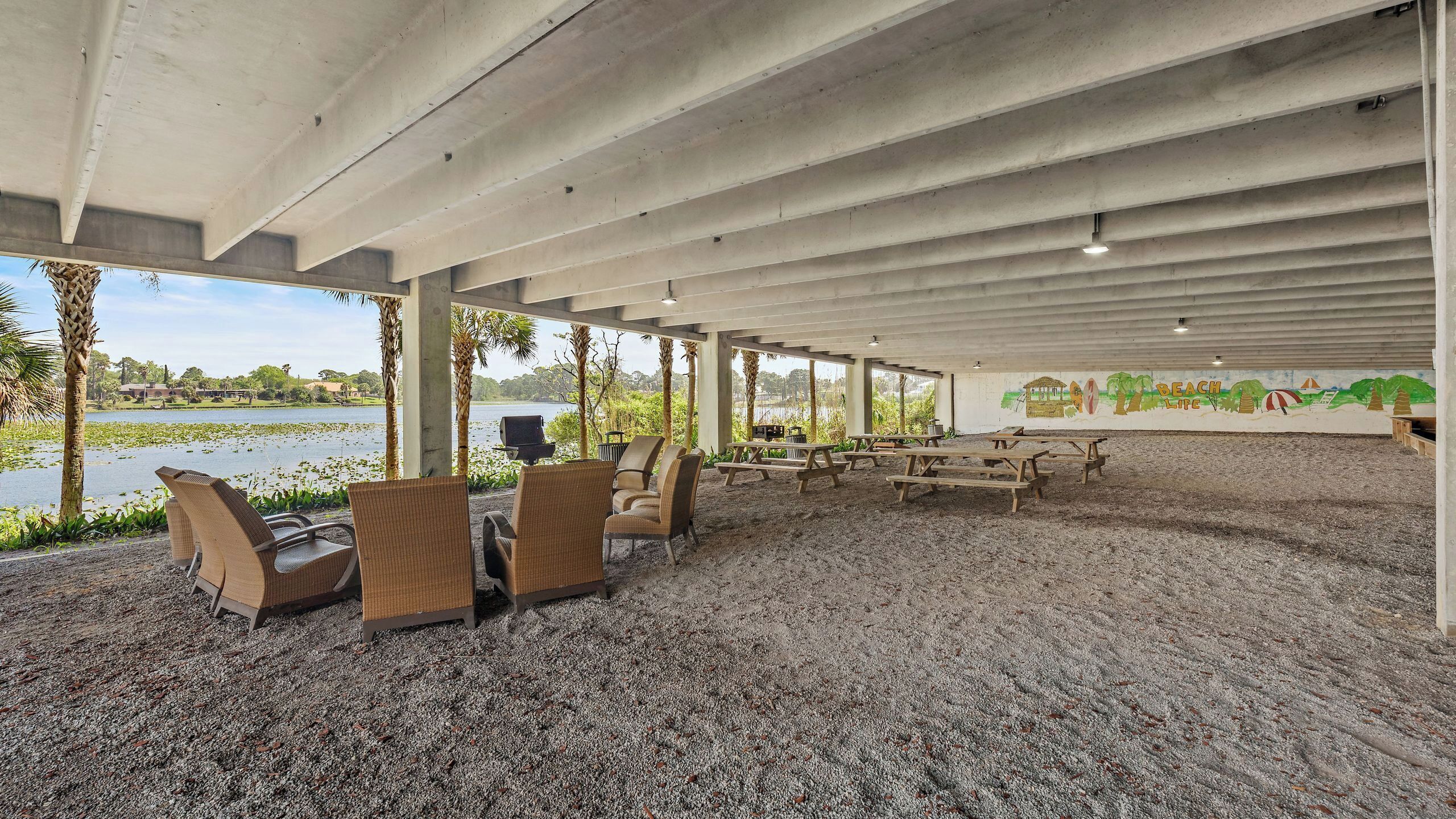 An outdoor covered area with picnic tables and chairs, a view of a lake, and a mural depicting a beach scene.