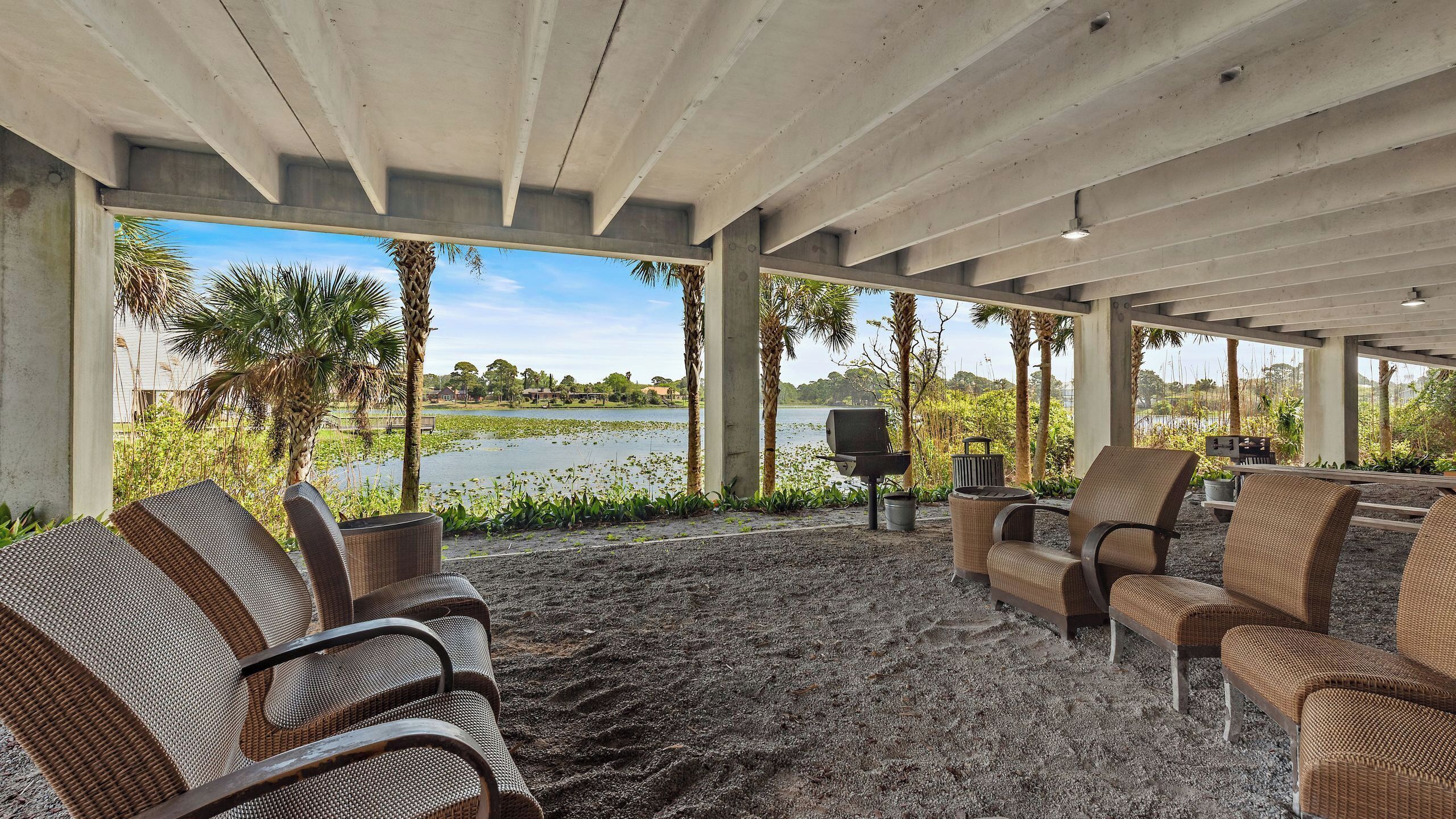 Un espacio exterior con mobiliario de mimbre y una parrilla, con vista a un lago con vegetación y casas al fondo.