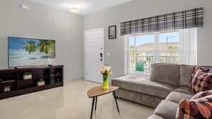 Ein Wohnzimmer mit Flachbildfernseher, einer Couch, einem kleinen Tisch mit einer Vase und einem Fenster mit Blick auf Gebäude.
