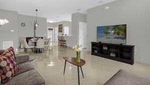 Ein modernes Wohnzimmer mit Flachbildfernseher, einem Essbereich mit Stühlen, einem Couchtisch mit Vase und einem Sofa mit gemustertem Kissen.