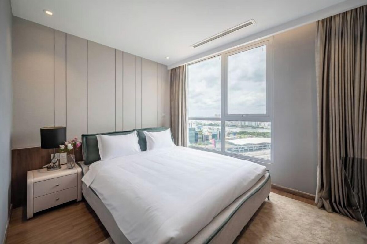 A hotel room with a large bed, a nightstand, a window with curtains, and a view of the city.