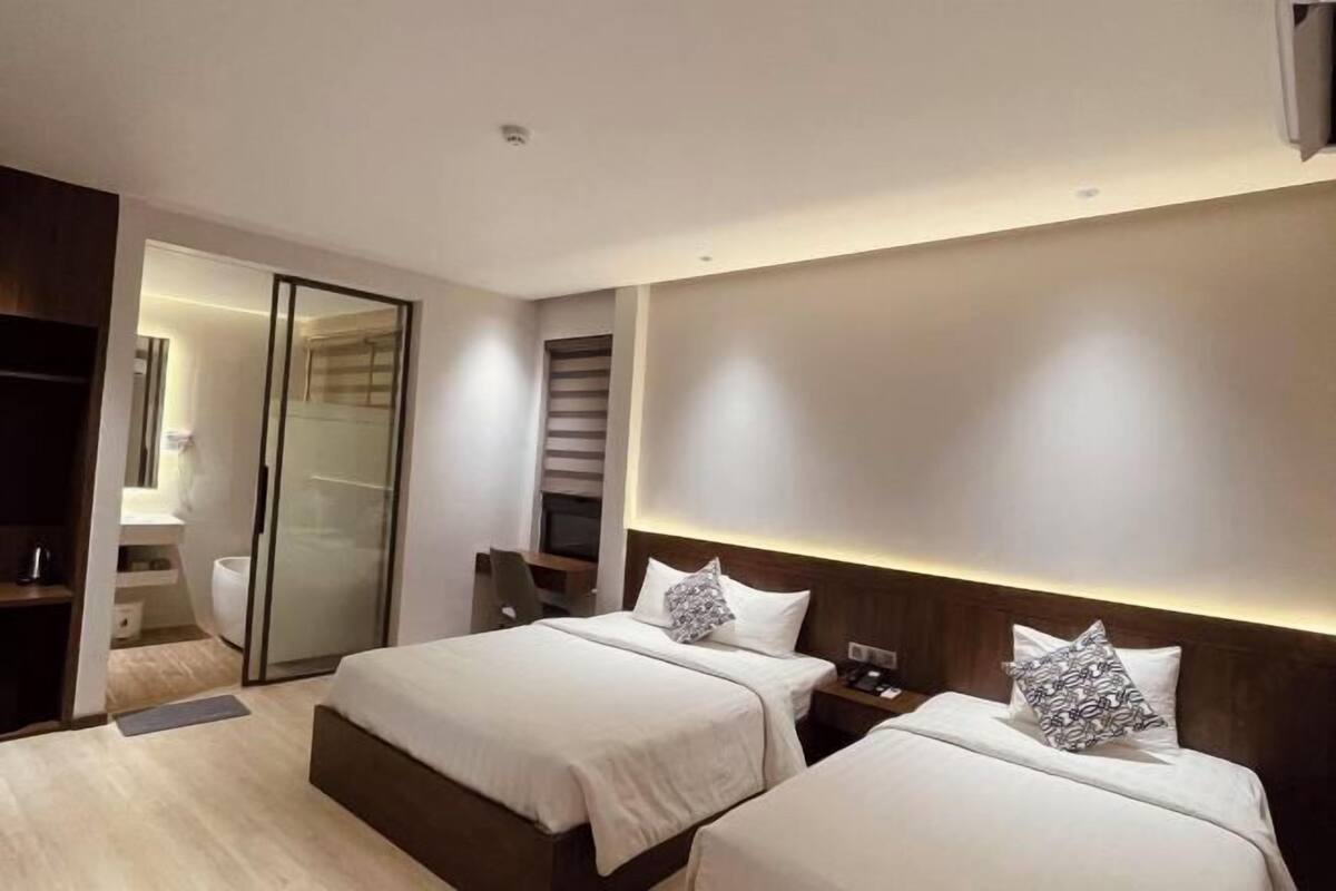 Une chambre d’hôtel moderne équipée de deux lits, d’un bureau, d’une chaise, d’une armoire et d’une salle de bain visible à travers une porte coulissante.