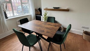 Uma área de jantar com mesa de madeira e cadeiras estofadas em verde, uma janela com vista para a rua, e uma estante com objetos decorativos.