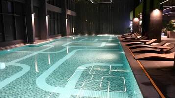 An indoor swimming pool with a tiled floor, lounge chairs, and a dark interior.