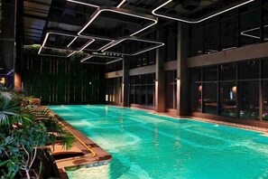 An indoor swimming pool with a view of the city at night, surrounded by plants and modern lighting fixtures.
