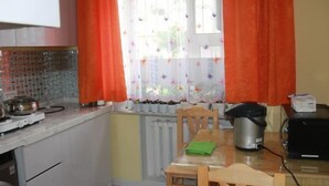 Una pequeña cocina con estufa, hervidor, cocedor de arroz y microondas. Hay una ventana con cortinas y un radiador debajo.