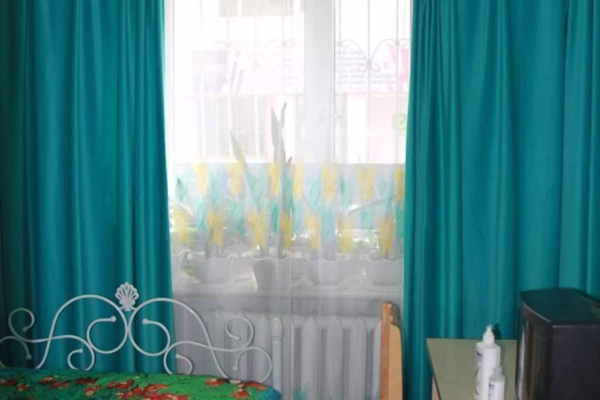A room with teal curtains, a bed with a floral bedspread, a radiator, and a small table with a bottle.
