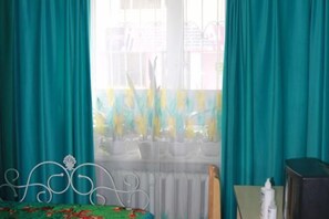 Ein Zimmer mit türkisblauen Vorhängen, einem Bett mit floralem Überwurf, einem Heizkörper und einem kleinen Tisch mit einer Flasche.