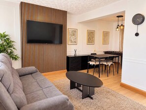 A modern living room with a flat-screen TV, a grey sofa, a coffee table, and a dining area with a table and chairs.