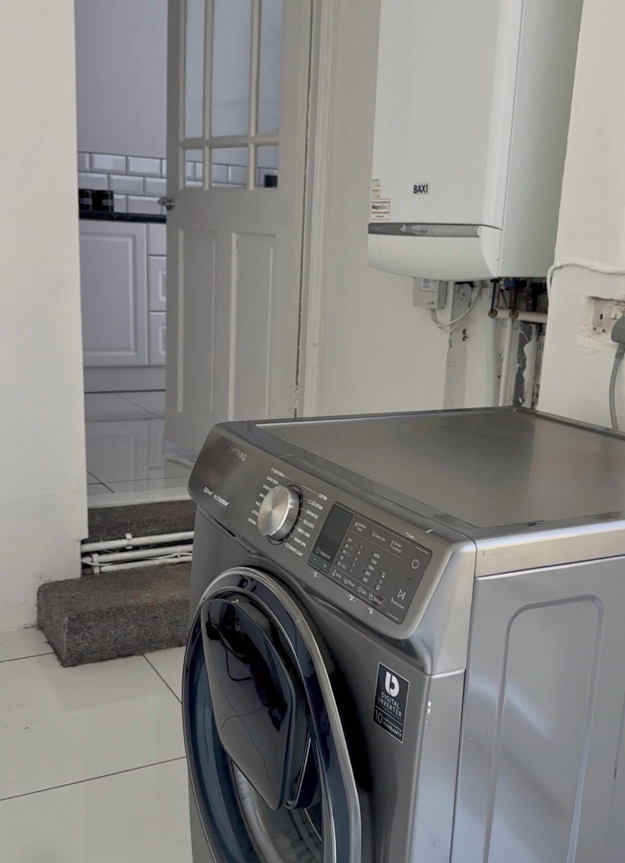A front-loading washing machine in a laundry room with a digital control panel, a door leading to a hallway, and a white wall-mounted water heater.
