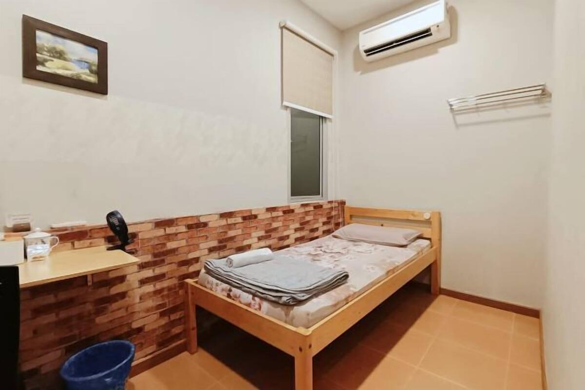 A small room with a wooden bed, a desk with a chair, a window, and an air conditioning unit.