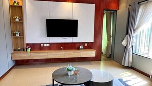 A modern living room with a flat-screen TV mounted on a wall, a wooden console table, a round coffee table, and a view of a green outdoor area. - TDH HOTEL (Ikeja)