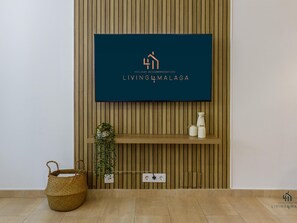 A flat-screen TV mounted on a wooden wall with a logo for 'Holiday Accommodation Living & Malaga'. Below the TV, there's a shelf with decorative items and a power outlet.