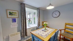 A dining area with a table set for two, a clock on the wall, and a window with curtains.