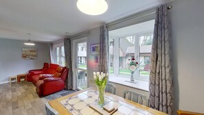 A spacious living room with a red sofa, a dining table with a vase of flowers, and large windows offering a view of the outside.